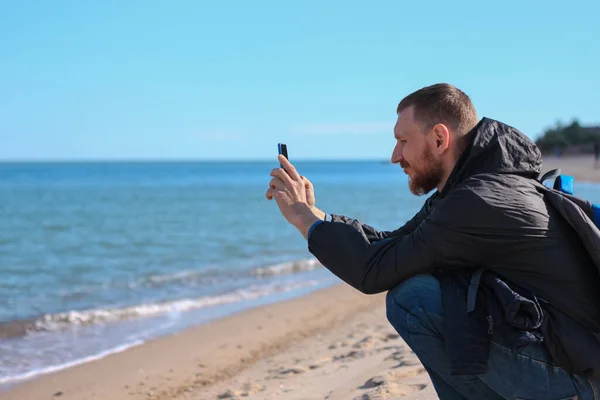 Bearded man in black raincoat on a blue sea landscape background with smartphone in hands talking. Middle aged guy calling friends on the cell phone on nature, people communication lifestyle concept.