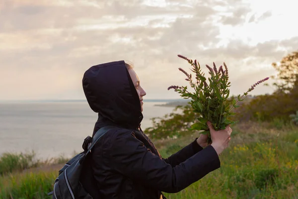 女孩穿着黑色雨衣 背包着野花 带着海景的自然背景 趋势超当地旅行 旅行者的家庭出游 回收概念 检疫结束 — 图库照片