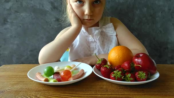 新鮮な果物やお菓子をテーブルの上に自宅で女の子 子供は健康的または有害なジャンクフードを選択するものを考えます 子供は自然夏の果実 ビタミンベジタリアンの食事を選択して楽しむ — ストック動画