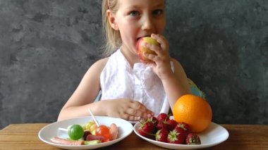 Evdeki masada taze meyve ve şekerle duran küçük kız. Çocuk sağlıklı ya da zararlı abur cubur seçmeyi düşünür. Çocuk doğal yaz meyvelerini, vitaminleri ve vejetaryen yemeklerini seçiyor..