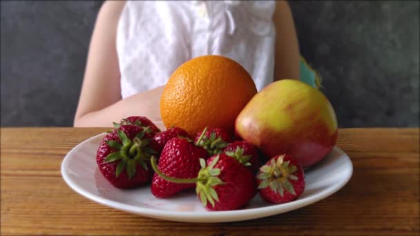 自宅のテーブルの上の白いプレートの上に新鮮な果物を持つ少女 子供は健康的な食事を選ぶ 子供は自然の夏の果実 ビタミンやミネラル ベジタリアンの食事アレルギー反応を食べることを楽しむ — ストック動画