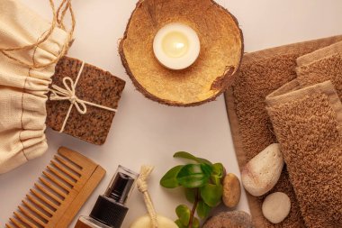 Spa ve sağlık konsepti, çevre çantasındaki doğal kahve sabunu, yağ kozmetik spreyi, soyulmuş kum taşı, havlu, ahşap saç tarağı. Bej rengi spa seti. Kahverengi pastel banyo aksesuarları ve ürünler üst görünümde.
