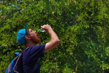 Genç adam elinde bir şişe su tutuyor ve içiyor. Kırmızı sakallı hippi, sırt çantalı mavi tişörtlü güneşli bir günde doğada duruyor. Spor koşusu, aktif yaşam tarzı, insan vücudu bakımı..
