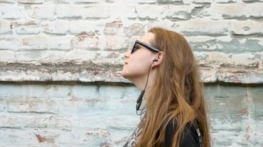 Siyah tişörtlü ve rock tarzında güneş gözlüklü genç çekici bir kız. Arka planda müzik dinliyor, kulaklık takıyor ve dans ediyor. Kablosuz kulaklıklı hippi eski tuğla duvarın yanında gülümsüyor..