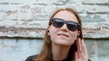 Siyah tişörtlü ve rock tarzında güneş gözlüklü genç çekici bir kız. Arka planda müzik dinliyor, kulaklık takıyor ve dans ediyor. Kablosuz kulaklıklı hippi eski tuğla duvarın yanında gülümsüyor..