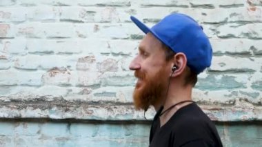 Siyah tişörtlü genç kızıl sakallı bir adamın portresi ve arka planda rock tarzında kulaklıkla müzik dinlerken. Kablosuz kulaklıklı hippi adam eski mavi tuğla duvar zemininde.