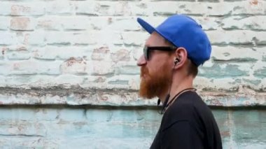 Siyah tişörtlü genç kızıl sakallı bir adamın portresi ve arka planda rock tarzında kulaklıkla müzik dinlerken. Kablosuz kulaklıklı hippi adam eski mavi tuğla duvar zemininde.