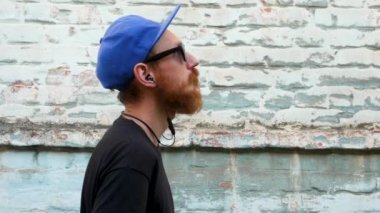 Siyah tişörtlü genç kızıl sakallı bir adamın portresi ve arka planda rock tarzında kulaklıkla müzik dinlerken. Kablosuz kulaklıklı hippi adam eski mavi tuğla duvar zemininde.