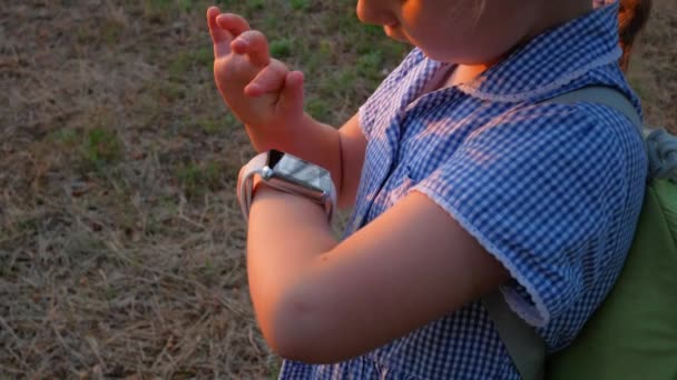 公園のスマートウォッチ屋外を使用して子供 子供はスマートフォンのビデオ通話で話しています 時計の閲覧インターネット上のタッチスクリーンディスプレイを使用して女子高生 Gpsトラッカー付きスマート腕時計 — ストック動画