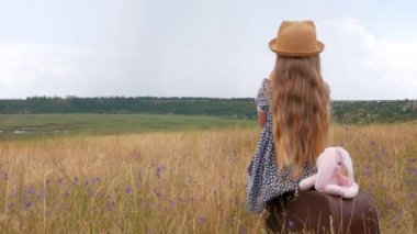 Uzun saçlı, hasır şapkalı ve elbiseli bir çocuk klasik bir bavula oturup fotoğraf çekiyor. Doğanın arka planına bakan yumuşak oyuncaklı tatlı bir çocuk. Macera seyahati konsepti
