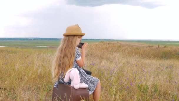 小女孩 头戴草帽 头戴长发 穿着长裙 坐在老式手提箱上拍照 可爱的孩子 带着柔软的玩具 看着大自然的蓝景背景 复古风格的冒险旅行概念 — 图库视频影像