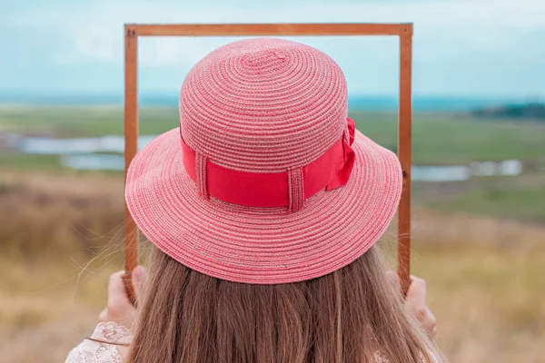戴着粉红草帽的女孩手里拿着空木框 在大自然的景观背景上默默无闻 一个长头发的女人的画像 在照片的背面 创意概念 — 图库照片