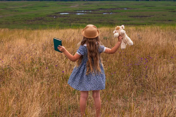 穿着草帽和衣服的小女孩呆在老式手提箱附近看书 可爱的孩子 带着柔软的狗玩具 手拿笔记本看着大自然的兰斯海景背景 冒险概念复古风格 — 图库照片