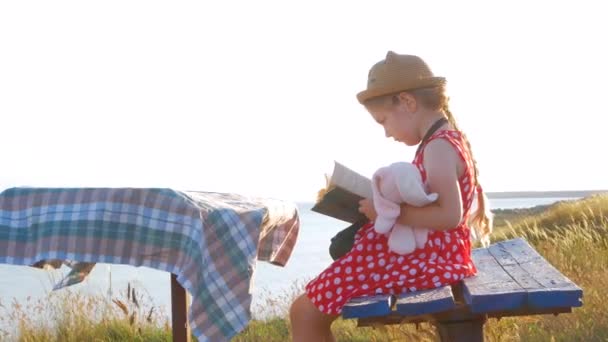 穿着草帽和衣服的小女孩坐在老式长椅上看书 可爱的孩子 带着柔软的兔子玩具 手里拿着笔记本 看着海景背景 友谊概念 — 图库视频影像