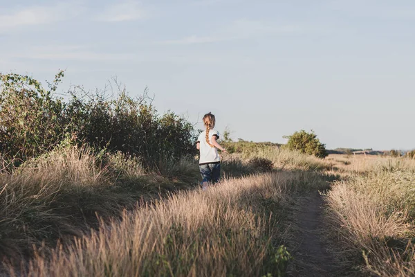 stock image Adorable little girl with long braid running down the hill back view. Happy child running in wild grass countryside landscape. Family walking on fresh air. Local hiking travel concept.