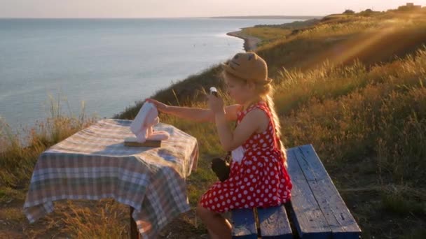 穿着草帽和衣服的小女孩坐在老式长椅上拍照 可爱的孩子 带着柔软的粉色兔子玩具 手里拿着笔记本 看着海景背景 友谊概念 — 图库视频影像