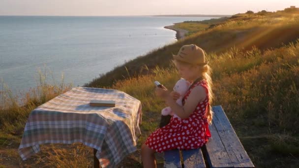 藁帽子の子供の女の子とドレスはヴィンテージベンチに座って本を読んでいます 海の自然景観を背景に手にノートを見て柔らかいウサギのおもちゃでかわいい子供 友情の概念 — ストック動画