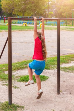 Yatay çubukta asılı duran kız. Çocuk hareket egzersizleri yapıyor ve dışarıda oyun oynuyor. Temiz havada sağlıklı bir spor aktivitesi. Mutlu bir çocukluk. Spor stadyumunda çocuklar için beden eğitimi.