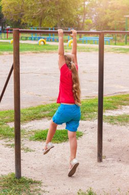 Yatay çubukta asılı duran kız. Çocuk hareket egzersizleri yapıyor ve dışarıda oyun oynuyor. Temiz havada sağlıklı bir spor aktivitesi. Mutlu bir çocukluk. Spor stadyumunda çocuklar için beden eğitimi.