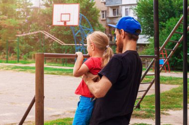 Yatay çubukta asılı duran kız. Babası olan bir çocuk dışarıda oyun parkında egzersiz yapıyor. Sağlıklı aile sporu aktivitesi. Mutlu bir çocukluk. Spor stadyumunda çocuklar için beden eğitimi.