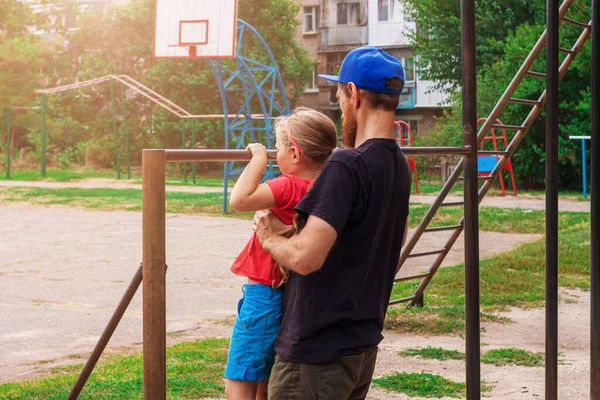 Yatay çubukta asılı duran kız. Babası olan bir çocuk dışarıda oyun parkında egzersiz yapıyor. Sağlıklı aile sporu aktivitesi. Mutlu bir çocukluk. Spor stadyumunda çocuklar için beden eğitimi.