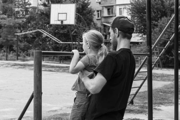 Girl hanging on horizontal bar. Child with father doing motion exercises on playground outdoor. Healthy family leisure sport activity. Kids physical training on sports stadium. Black and white photo. — Stock Image Girl Hanging Horizontal Bar Child Father Doing Motion Exercises Playground Stock Photo