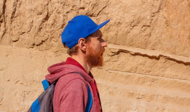 Sırt çantalı çekici bir yürüyüşçü dağ yolunda ilerliyor. Gezgin adam güneşli bir günde kil kaya arka planına bakıyor. Macera seyahati ve keşif..