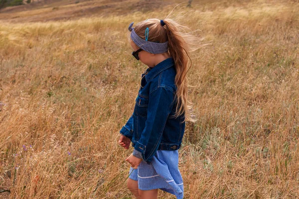 漂亮的小女孩穿着牛仔夹克 戴着黑色太阳镜 穿着蓝色格子呢裙 穿着黄色的草地 在乡村风景中快乐而时尚的金发孩子 可爱的孩子走在户外的乡村道路上 — 图库照片