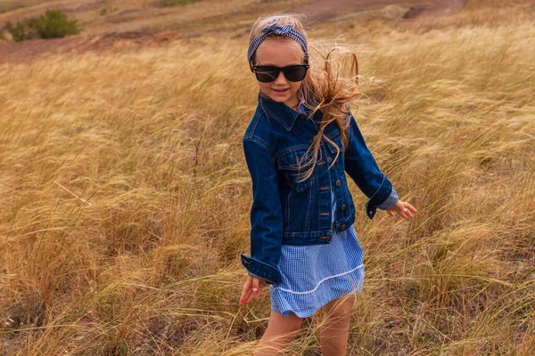 Kot ceketli, siyah güneş gözlüklü, sarı çim tarlasında mavi ekose elbiseli sevimli küçük kız. Kırsal alanda uzun sarı saçlı mutlu bir çocuk. Kırsal alanda gezen sevimli bir çocuk..