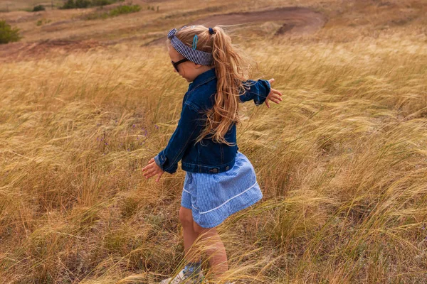 Kot ceketli, siyah güneş gözlüklü, sarı çim tarlasında mavi ekose elbiseli sevimli küçük kız. Kırsal alanda uzun sarı saçlı mutlu bir çocuk. Kırsal alanda gezen sevimli bir çocuk..