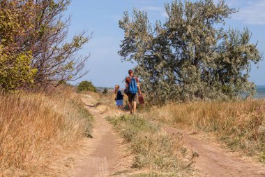 Bavullu baba ve kızı doğa manzarası üzerinde off-road yürüyorlar. Açık havada babasıyla kucaklaşan küçük sarışın kız. Sevgi dolu çocuk babasını kucaklıyor. Babalar Günü aile tebrik kartı..