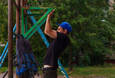 Sporcu, barfiks çekiyor. Erkek sporcu, teneffüste dışarıda oyun alanında spor yapıyor. Sırt çantalı sakallı genç adam iş gününden sonra ısınıyor. Etkin yaşam biçimi.