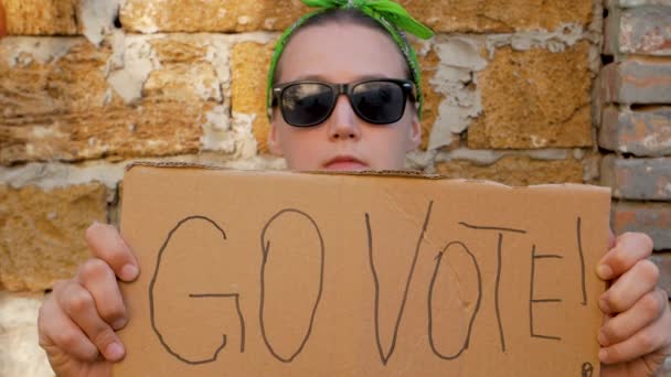女の子はゴー投票で段ボールを示しています レンガの壁の都市の背景にサイン 投票の概念 政治的な選択をし 声を使う 黒いサングラスの女性は大統領選挙に行くために呼び出します — ストック動画