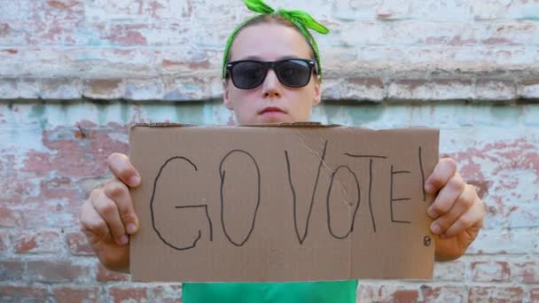 女孩展示硬纸板与去投票 在砖墙上签名城市背景 表决概念 做出政治选择 用你的声音 戴黑太阳镜的妇女呼吁去参加总统选举 — 图库视频影像
