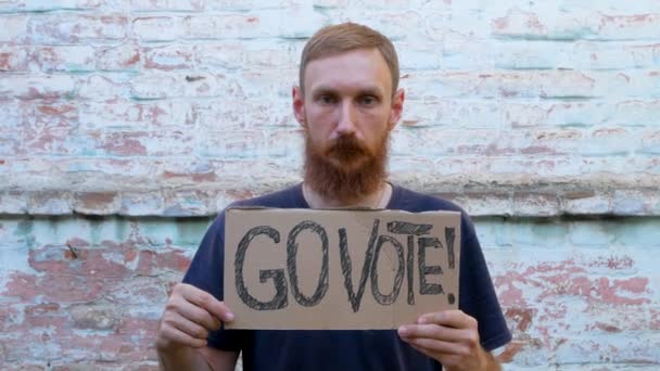 男人在砖墙的城市背景上展示带有Go Vote标志的硬纸板 表决概念 做出政治选择 用你的声音 大胡子的家伙打电话去参加总统选举 — 图库视频影像