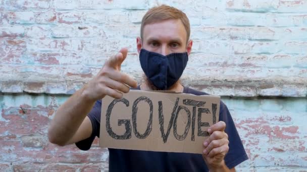 男はレンガの壁の都市の背景にゴー投票記号で段ボールを示しています 投票の概念 政治的な選択をし 声を使う ひげを生やした男が大統領選挙に行くことを要求する — ストック動画