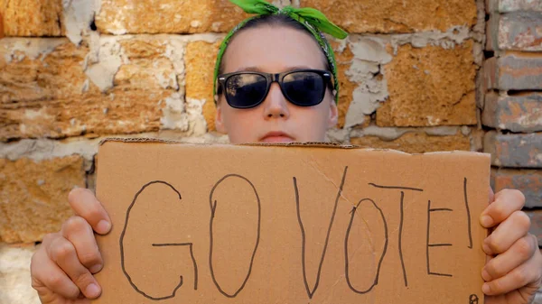 Girl shows cardboard with Go Vote sign on brick wall urban background. Voting concept. Make the political choice, use your voice. Woman in black sunglasses calls to go to the presidential elections.