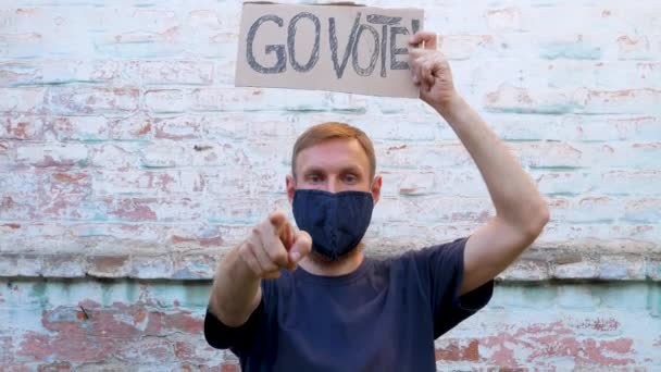 男はレンガの壁の都市の背景にゴー投票記号で段ボールを示しています 投票の概念 政治的な選択をし 声を使う 保護面マスクの男は大統領選挙に行くために呼び出します あなたの投票数 — ストック動画