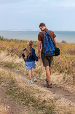 Baba ve kızı, doğa manzaralı bir tepenin üzerinde duruyor. Küçük sarışın kız babasıyla dışarıda kucaklaşıyor. Sevgi dolu çocuk babasını kucaklıyor. Babalar Günü aile tebrik kartı..