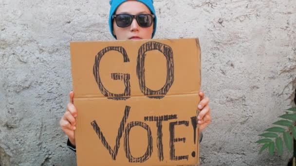 妇女在砖墙的城市背景上展示带有Go Vote标志的纸板 表决概念 做出政治选择 用你的声音 戴蓝帽的年轻女子应邀参加总统选举 — 图库视频影像
