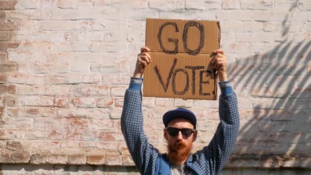 男人在砖墙的城市背景上展示带有Go Vote标志的硬纸板 表决概念 做出政治选择 用你的声音 戴著太阳镜的留胡子的家伙应邀参加总统选举 — 图库视频影像