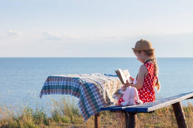 Şapkalı ve benekli elbiseli çocuk klasik bankta oturmuş kitap okuyor. Çocuk kutsal kitaba bakıyor ve deniz manzarası arka planında dua ediyor. Dostluk barış din umut kavramı.