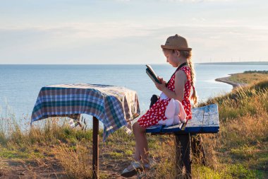 Şapkalı ve benekli elbiseli çocuk klasik bankta oturmuş kitap okuyor. Çocuk kutsal kitaba bakıyor ve deniz manzarası arka planında dua ediyor. Dostluk barış din umut kavramı.