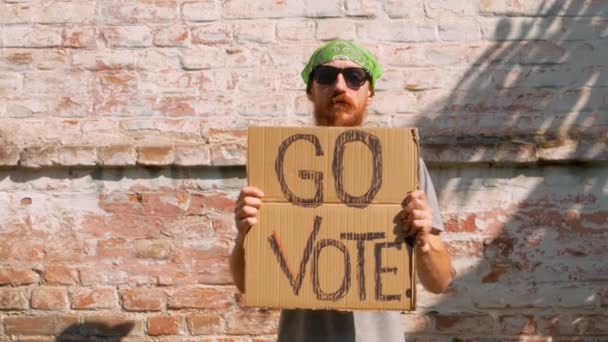 男人在砖墙的城市背景上展示带有Go Vote标志的硬纸板 表决概念 做出政治选择 用你的声音 头戴绿色头巾的大胡子的家伙邀请去参加总统选举 — 图库视频影像