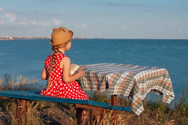 Şapkalı ve benekli elbiseli çocuk klasik bankta oturmuş kitap okuyor. Çocuk kutsal kitaba bakıyor ve deniz manzarası arka planında dua ediyor. Dostluk barış din umut kavramı.