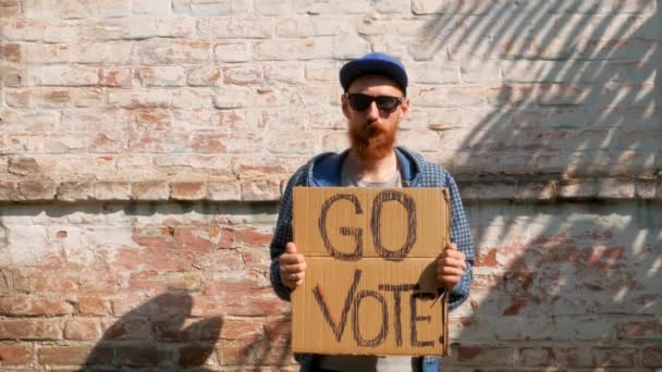男人在砖墙的城市背景上展示带有Go Vote标志的硬纸板 表决概念 做出政治选择 用你的声音 戴著太阳镜的留胡子的家伙应邀参加总统选举 — 图库视频影像