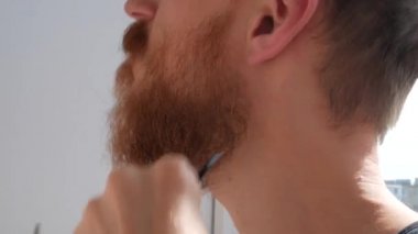 Yetişkin bir adam bıyığını ve sakalını makas ve tarakla kesiyor. Kafkas kızıl sakallı erkek evde saçlarını kesiyor. Kendi kendine saç kesimini kendin yap. Karantina karantinası sırasında özel bakım yap.