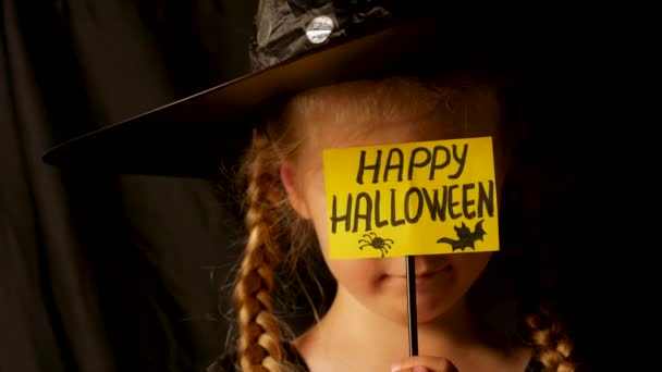 黒いウィザードの帽子の小さなかわいい女の子の魔女は挨拶を保持していますハッピーハロウィンカードはブーと笑いを言います 幽霊パーティーの衣装で身に着けている子供は怖いですカメラ4Kお祝いの秋の休日の映像 — ストック動画