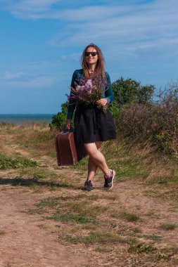 Uzun kahverengi saçlı, kot ceketli, siyah etekli, klasik valizli, mor çiçekli, off-road deniz manzaralı genç bir kadın. Taşra gezisinde otostopçu. Hayat tarzı fotoğrafçı kadın dışarıda dolaşıyor.
