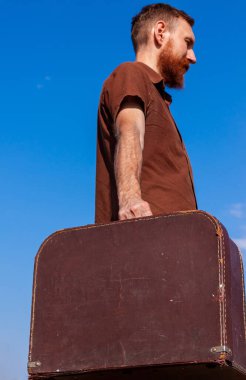 Yakışıklı hippi bıyıklı yakışıklı adam mavi gökyüzü arka planında kahverengi klasik bavul tutuyor. Dışarıda bagajı olan kızıl sakallı adam. Ciddi bir Kafkas gezgini. Vahşi erkek bakış açısı..